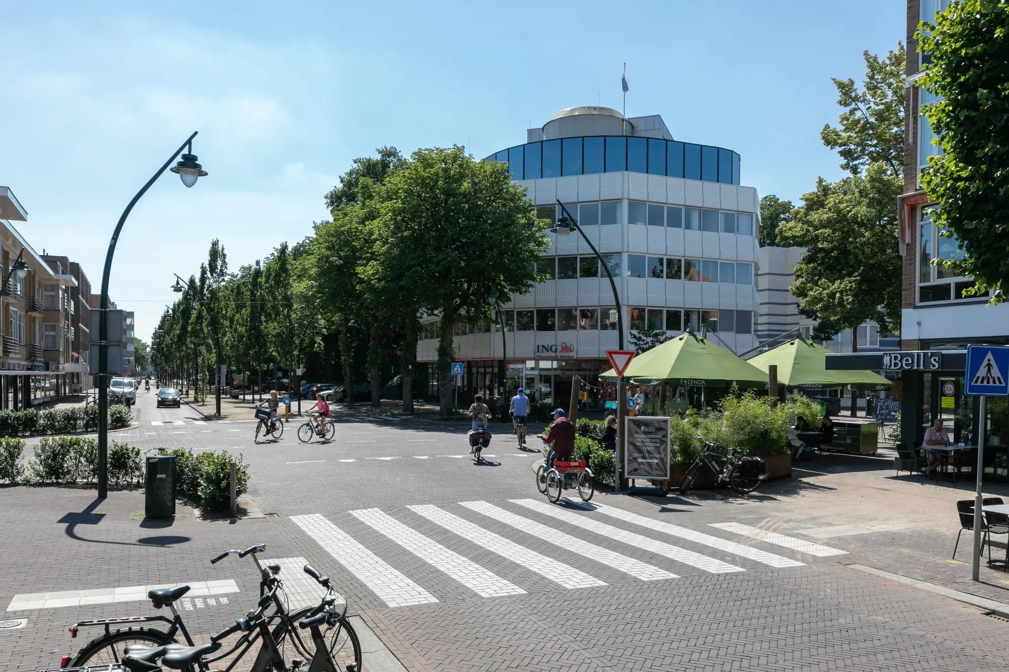 Straatbeeld van de 1e Hogeweg in Zeist met fietsers, voetgangers, een ING-gebouw en terrassen op een zonnige dag.